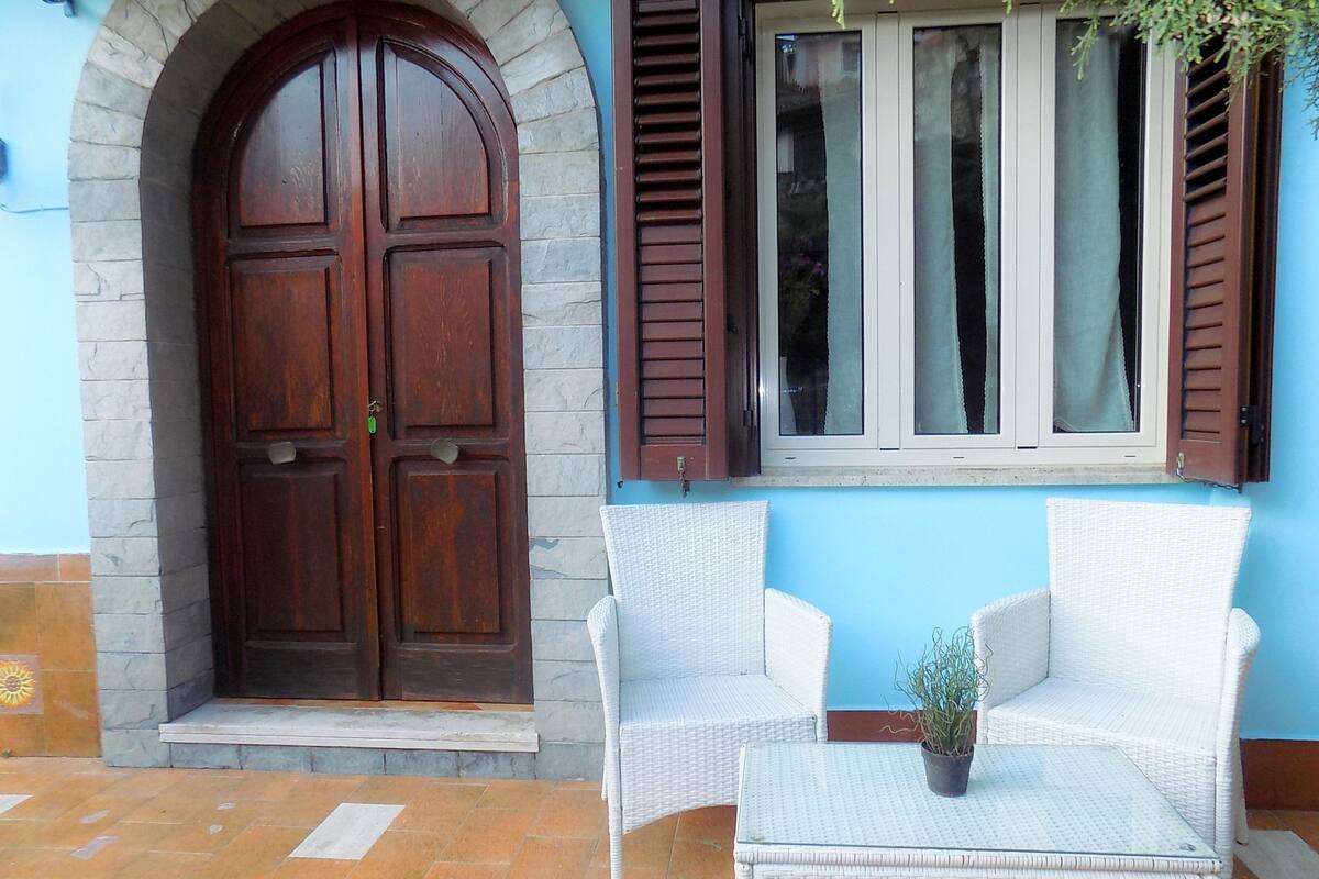 A building entrance with a large wooden door, a window with shutters, and two wicker chairs outside.
