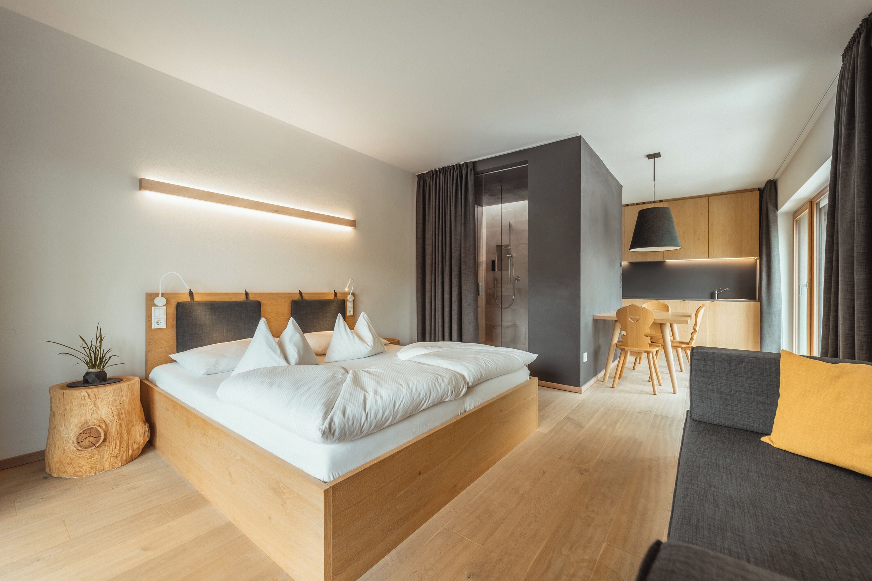 Habitación de hotel moderna con una cama grande, cabecera de madera, una mesita de noche con una planta, un sofá, una mesa de comedor con sillas y una zona de cocina con una lámpara colgante.