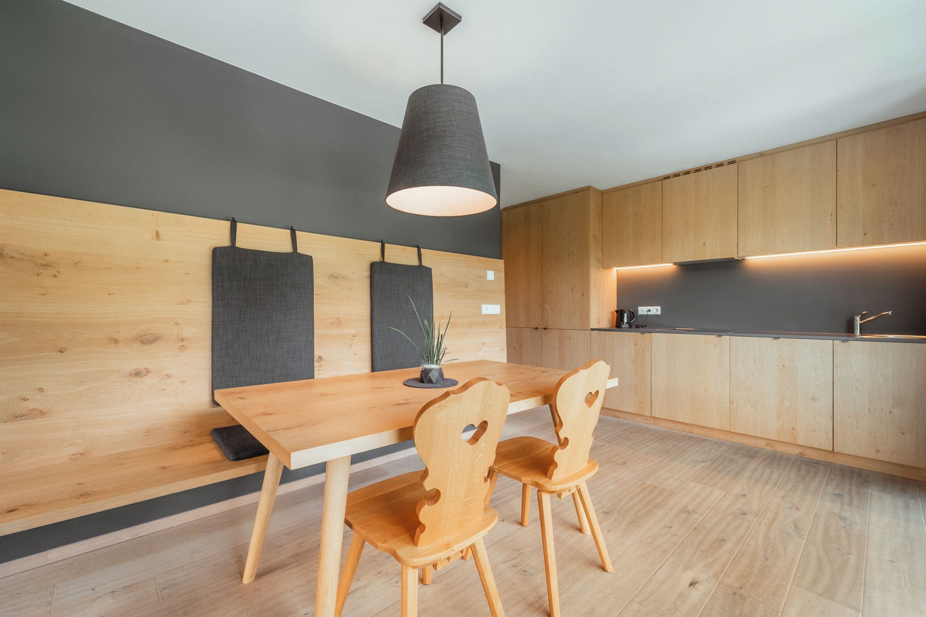 Una cucina moderna con mobili in legno, soffitto scuro e lampada a sospensione sopra un tavolo in legno con due sedie.