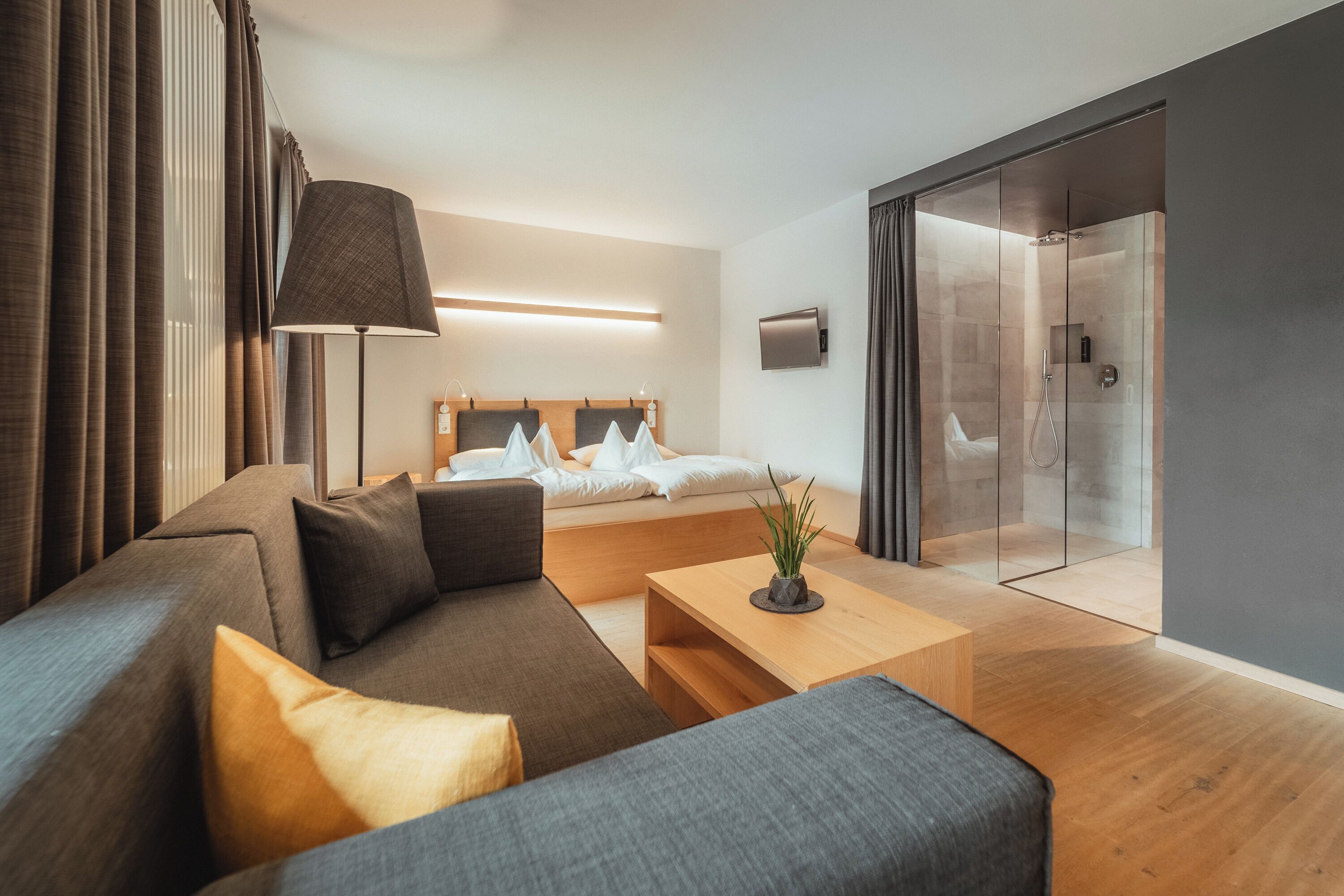 Una moderna camera d'albergo con divano, un letto con lenzuola bianche, un comodino, una lampada e una porta scorrevole in vetro che dà su uno spazio doccia.