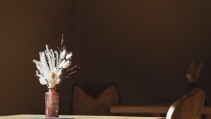 Una mesa de comedor con un jarrón de flores secas, una silla de madera y una pata de mesa de madera.