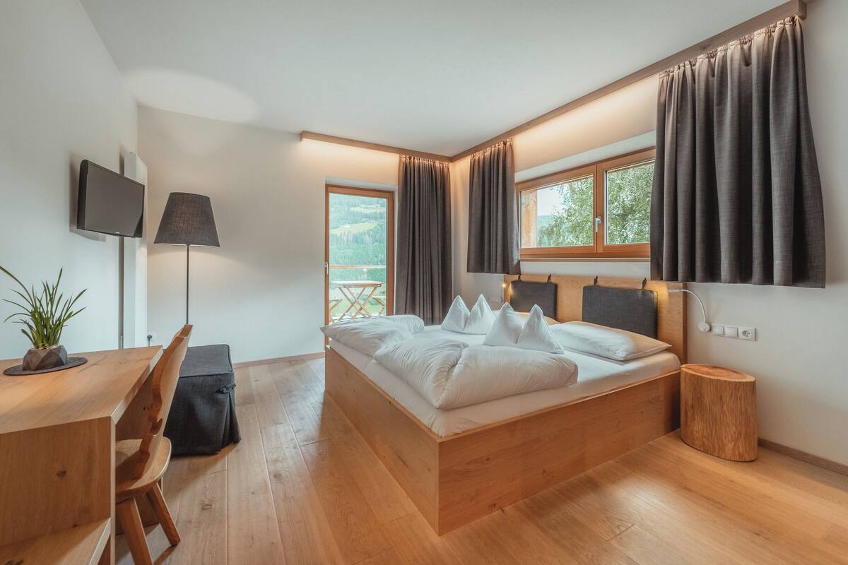 A modern bedroom with a wooden bed, a desk with a chair, a TV mounted on the wall, and a window with curtains.
