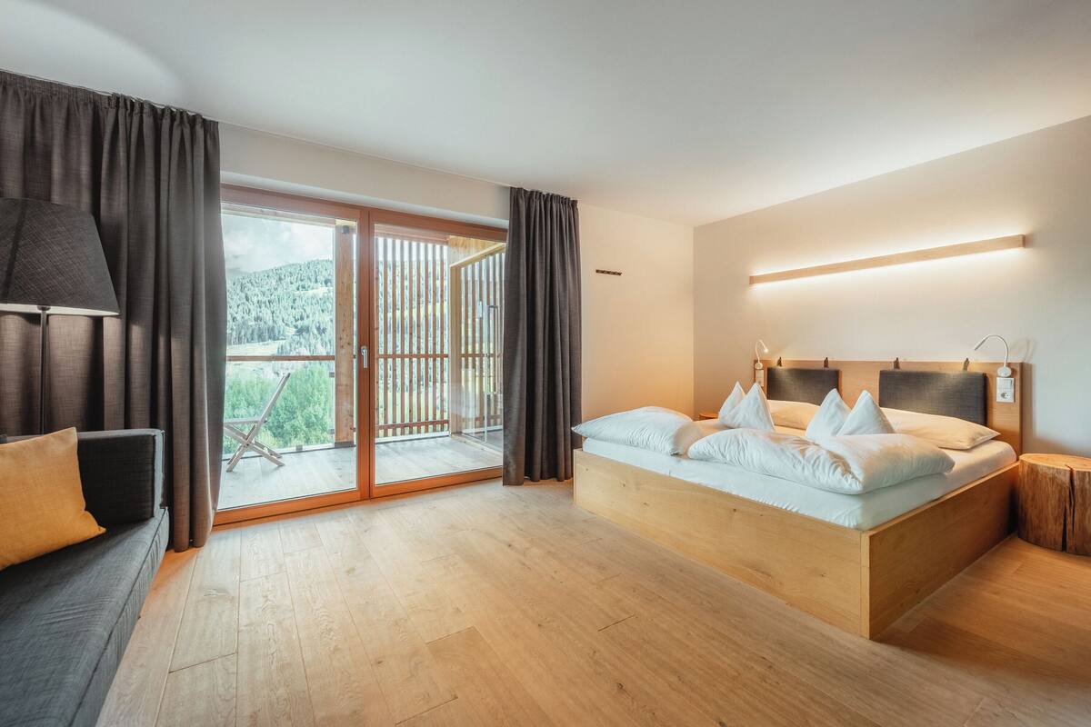 A modern bedroom with a wooden bed, a sofa, and a sliding glass door leading to a balcony.