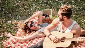 Una pareja recostada sobre una manta a cuadros en un área cubierta de hierba, con una cesta de picnic que contiene frutas y una guitarra.