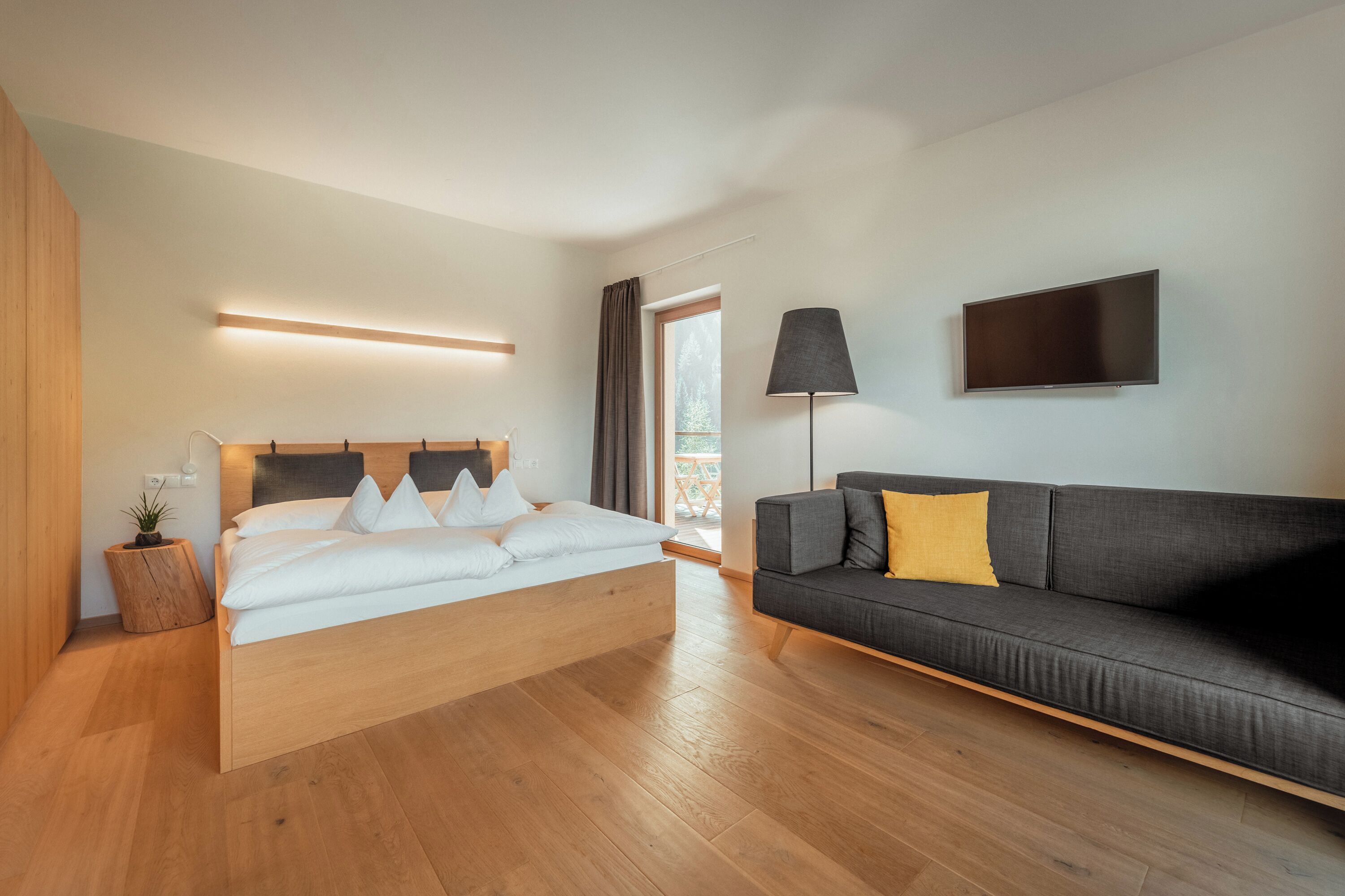 Ein modernes Schlafzimmer mit einem hölzernen Bett, einem grauen Sofa, einem Flachbildfernseher und einer Nachttischlampe.