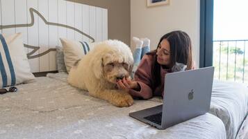 Una persona acostada en una cama con una computadora portátil, un perro blanco y esponjoso, y un cabecero con un diseño geométrico.