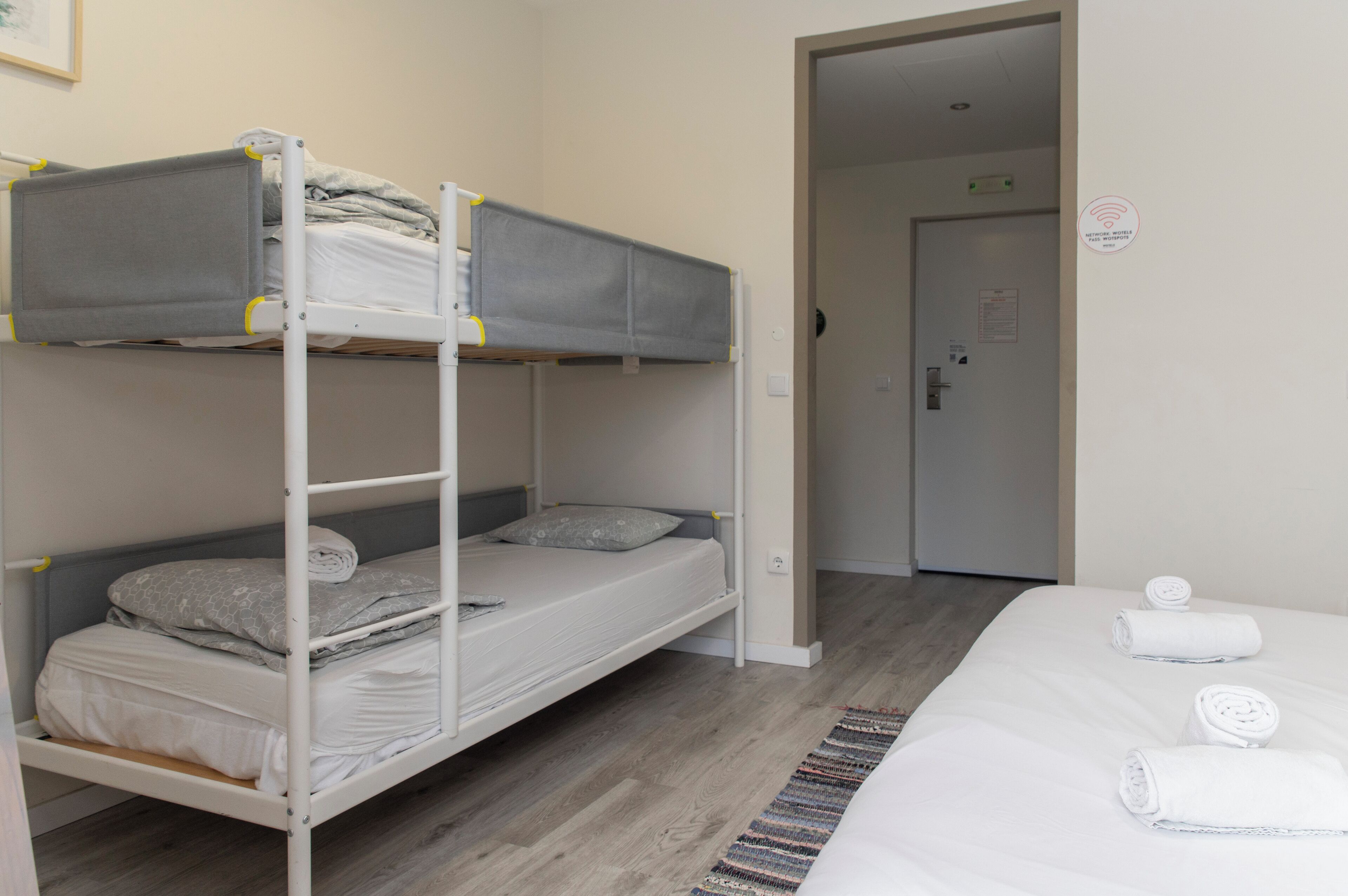 Habitación con literas, piso de madera, una cama blanca con toallas enrolladas y una puerta al fondo.