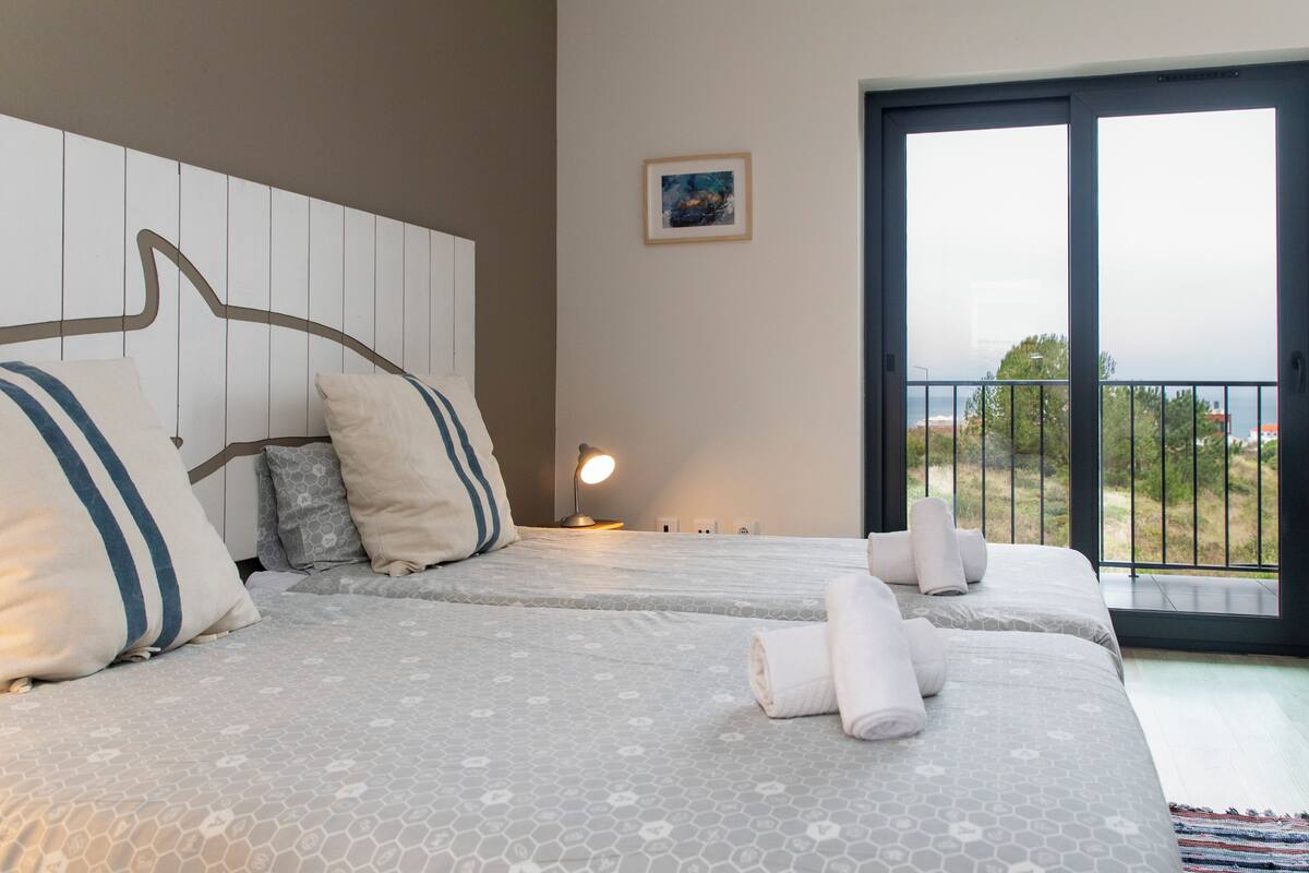 Habitación de hotel con dos camas, un ventanal amplio con vistas, cabecera de diseño original y mesita de noche pequeña con lámpara.