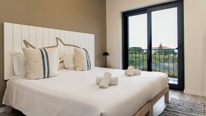 Une chambre à coucher avec un grand lit, une tête de lit, une table de chevet, une lampe et une vue sur l’extérieur, accessible par une porte coulissante.