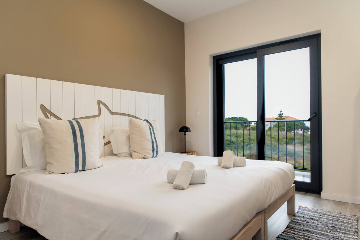Un dormitorio con una cama grande, cabecera, mesita de noche, lámpara y vistas al exterior a través de una puerta corredera.