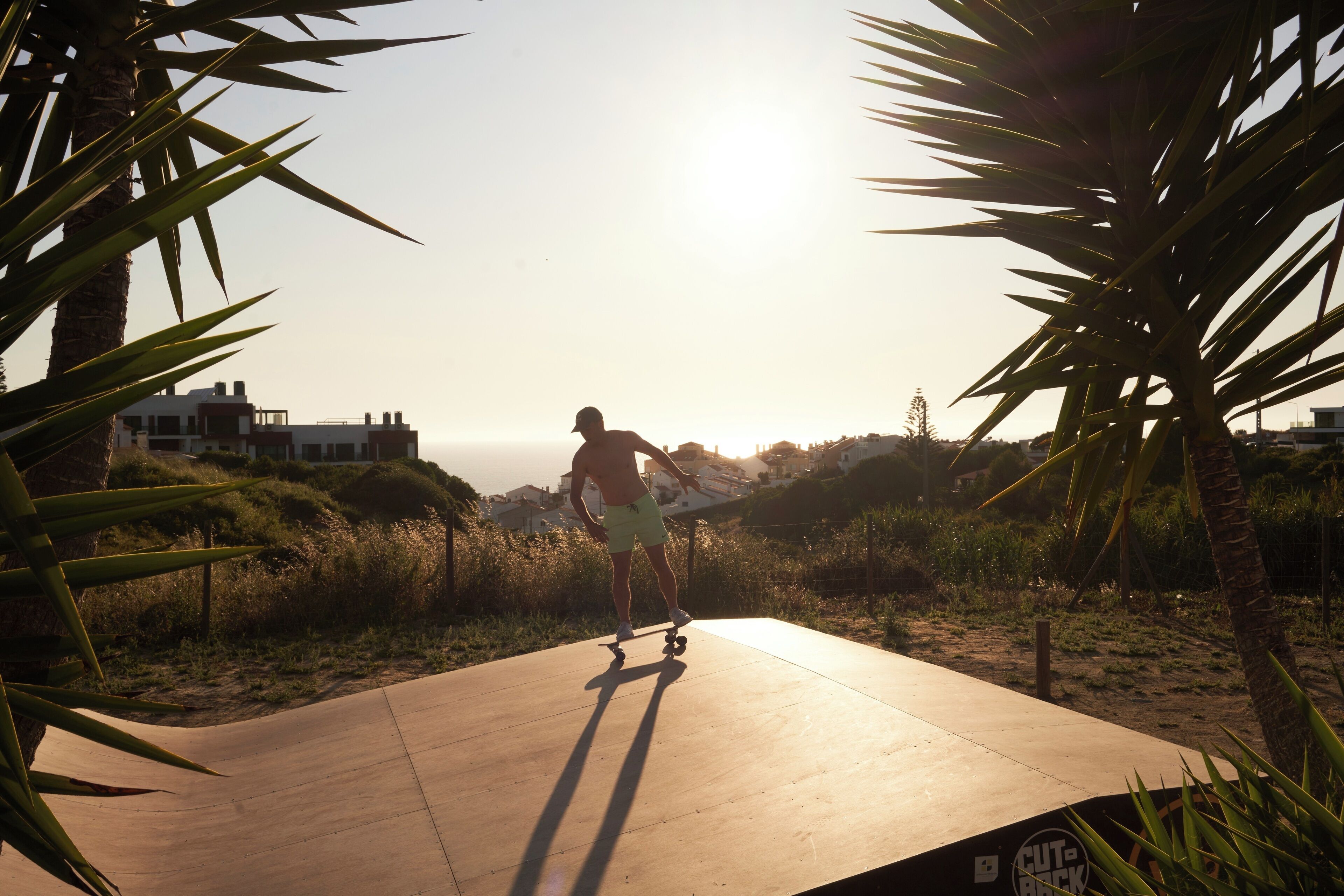 Une personne fait du skateboard sur une surface plane en béton, avec des palmiers et des bâtiments en arrière-plan.