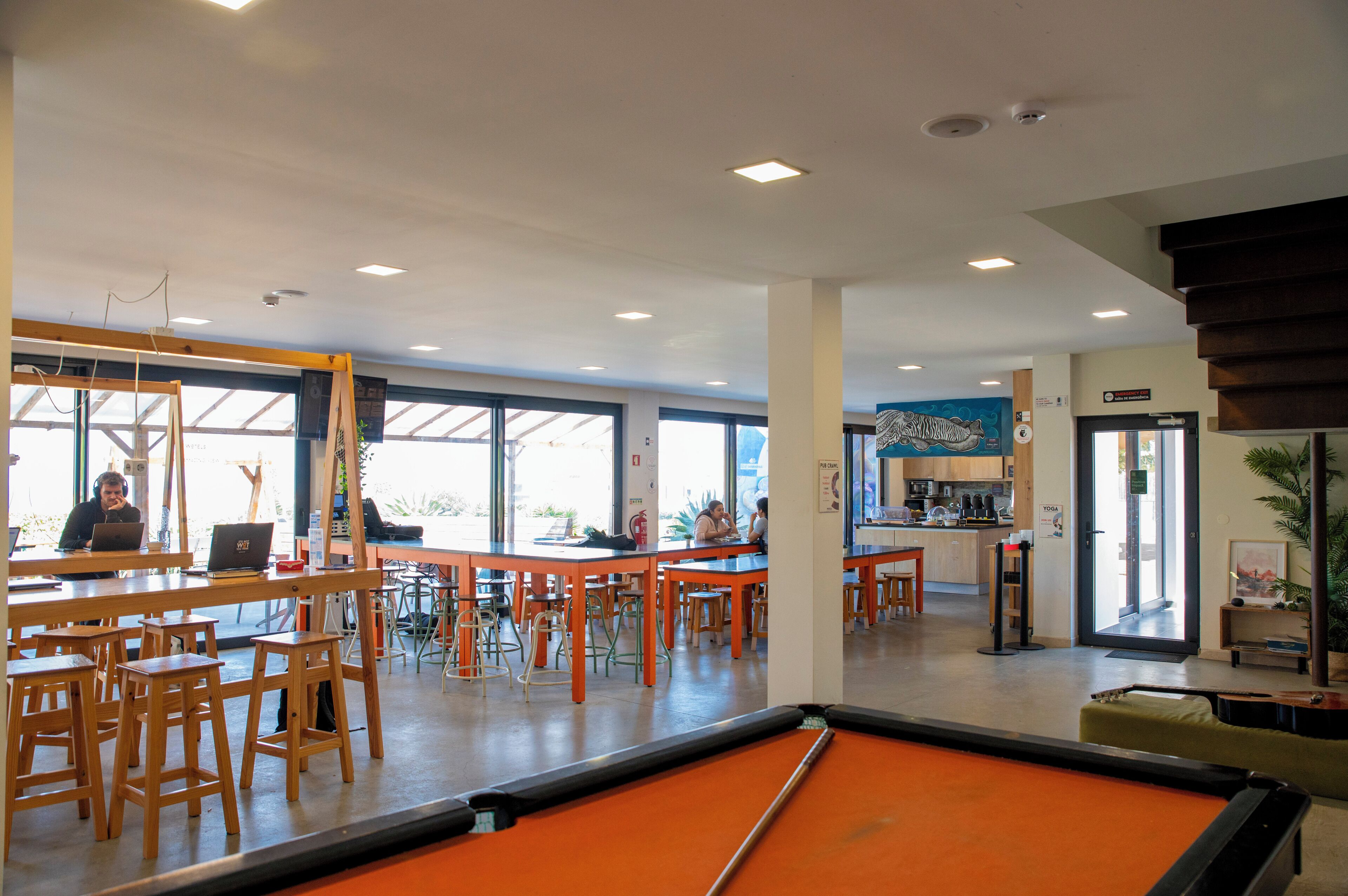 A pool table in a brightly lit indoor space with multiple seating areas, a bar, and large windows allowing natural light.
