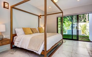 A four-poster bed with a canopy, a bedside table with a lamp, and a view of greenery outside.