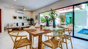 A dining area with a wooden table and wicker chairs, a sofa with cushions, and a view of a pool through large glass doors.