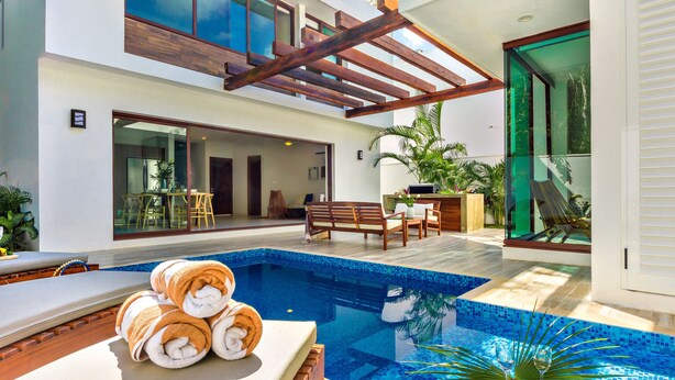 A modern outdoor pool area with a wooden deck, lounge chairs, and a glass-enclosed patio.