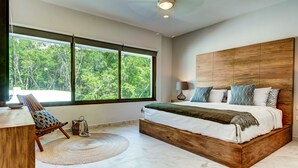 A bedroom with a large bed, a wooden headboard, a view of trees through the window, a chair, and a bedside table.