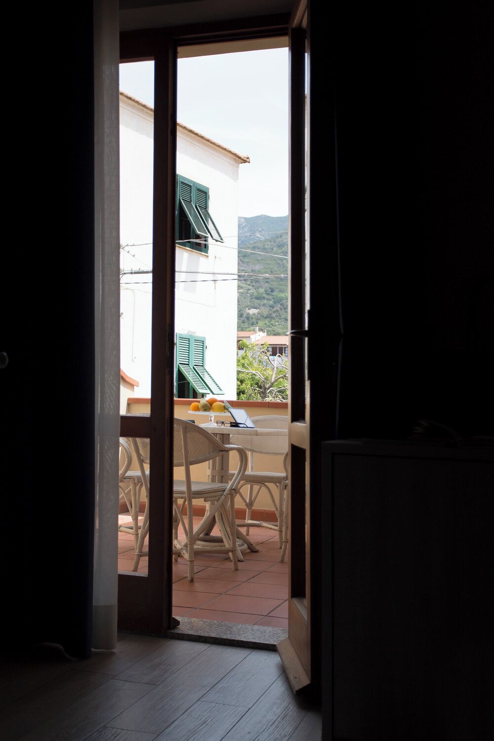 Une porte ouverte donnant sur un balcon où se trouvent une table et des chaises, un ordinateur portable et un panier de fruits.
