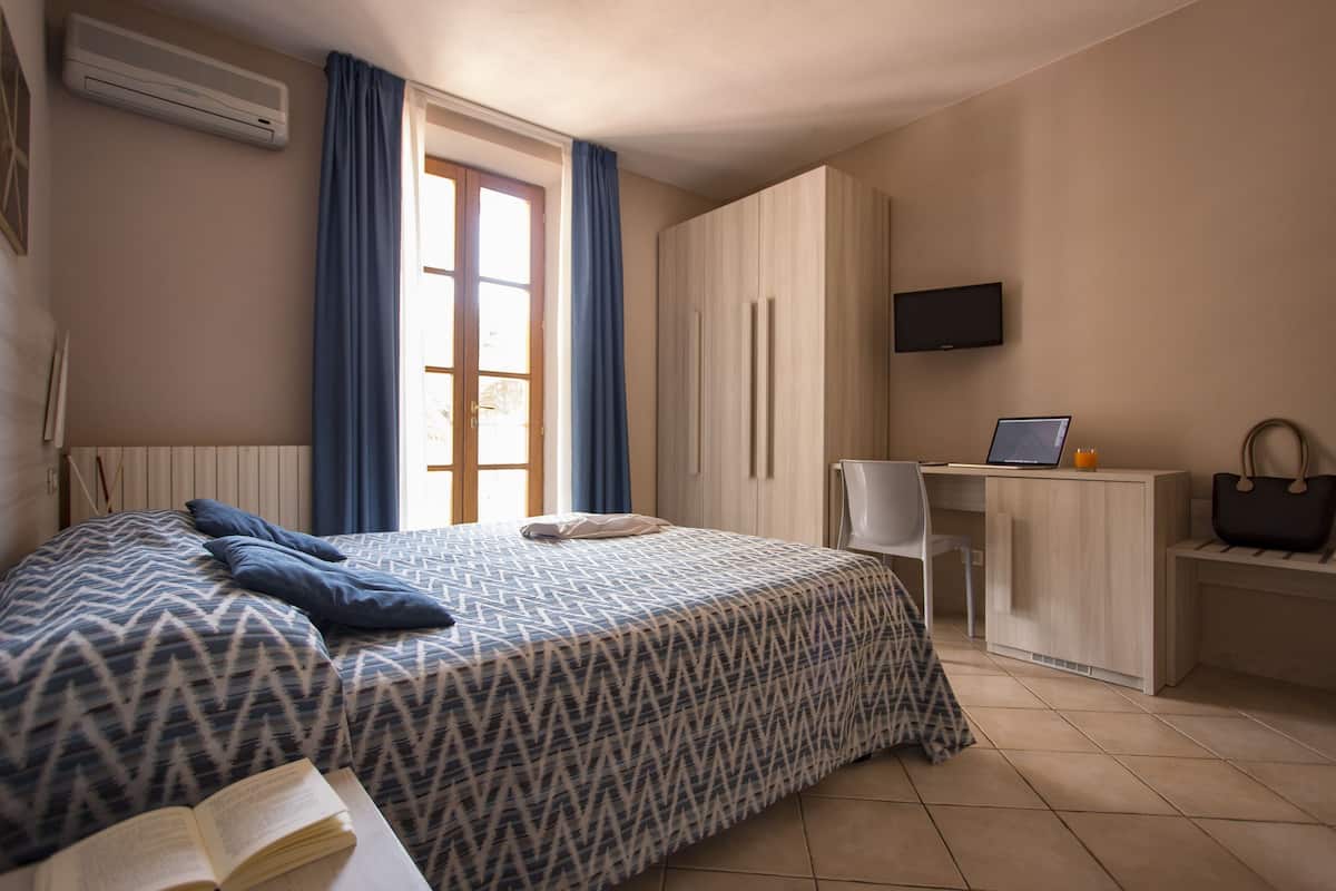 A bedroom with a bed, a desk, a chair, a TV, and a window with curtains.