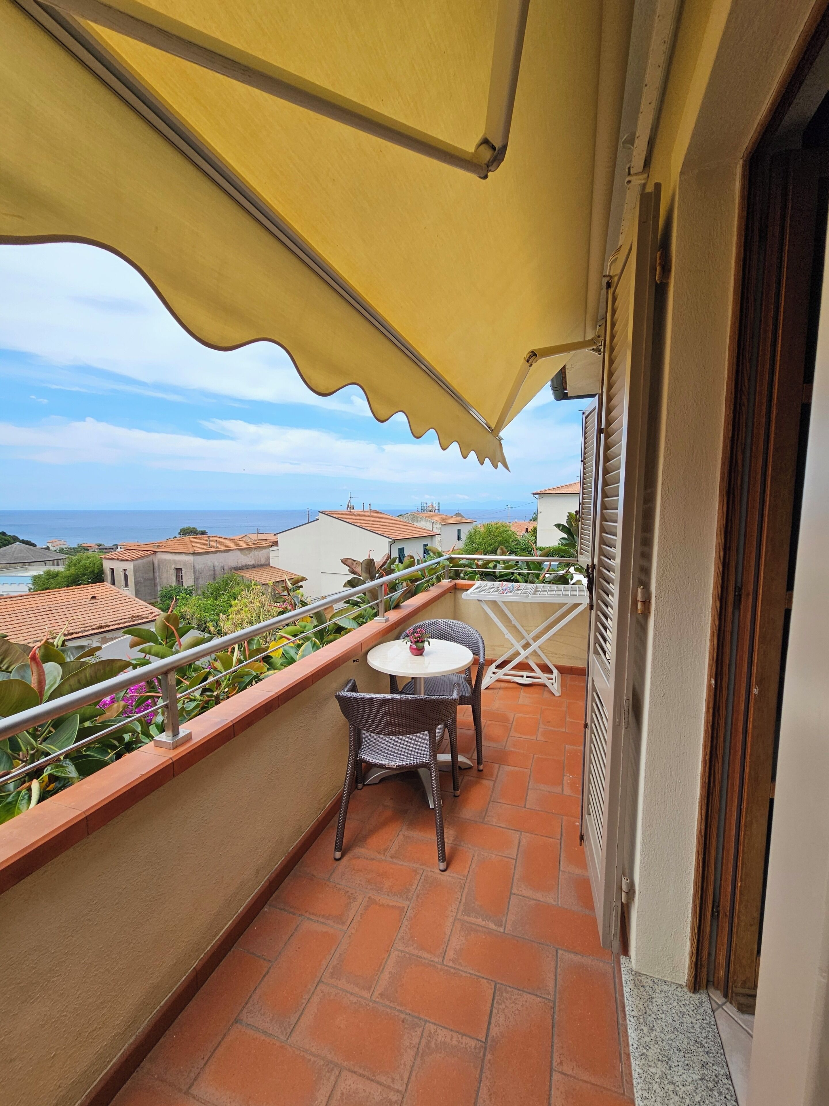 A balcony with a table and chairs, a yellow awning, and a view of the sea and buildings.