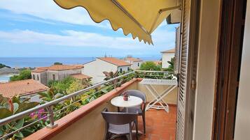 A balcony with a table and chairs, a yellow awning, and a view of the sea and buildings.