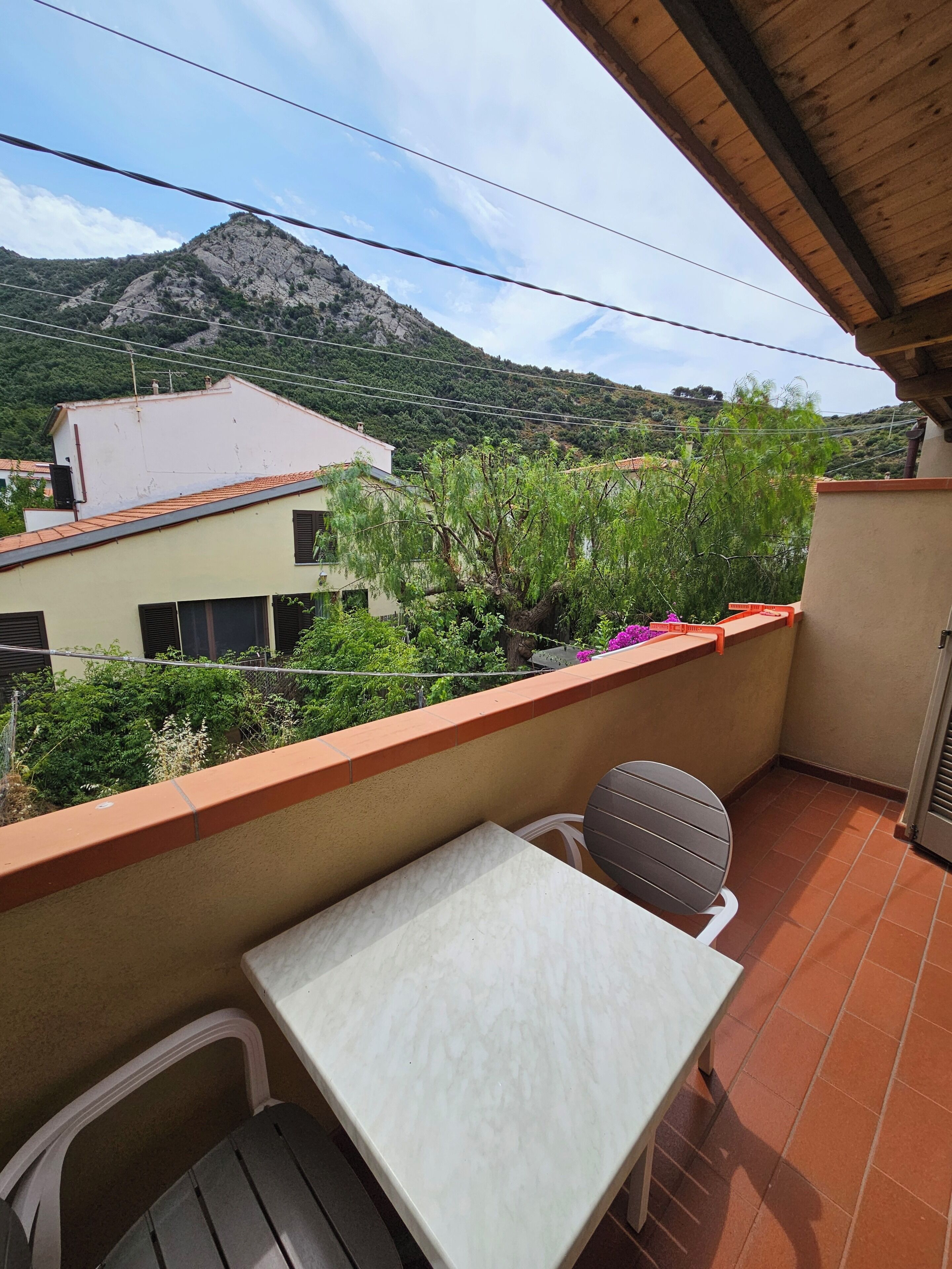 Un balcón con mesa y sillas, con vistas a un paisaje montañoso con vegetación y edificios.
