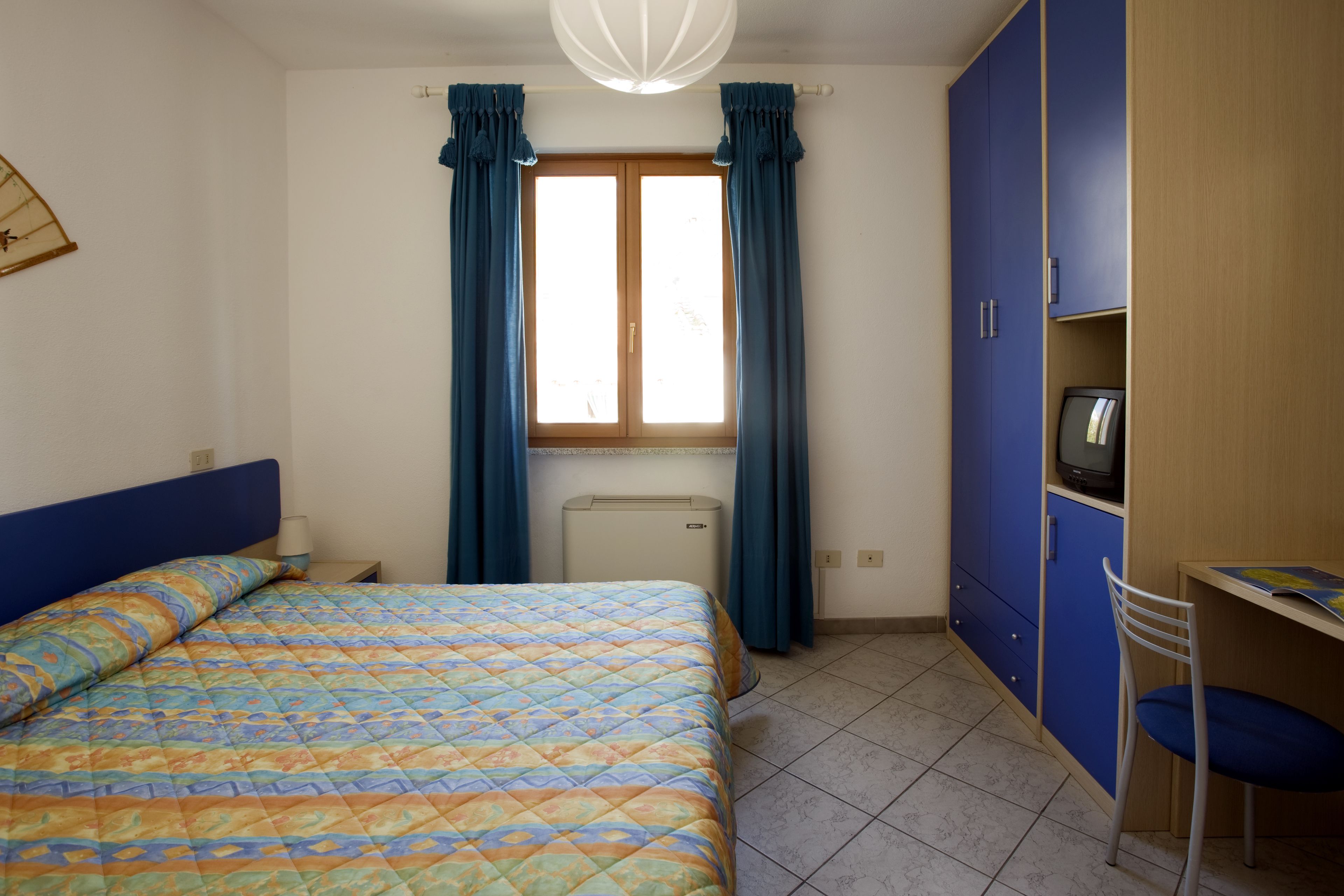 Quarto com cama, escrivaninha, cadeira, televisão e guarda-roupa.