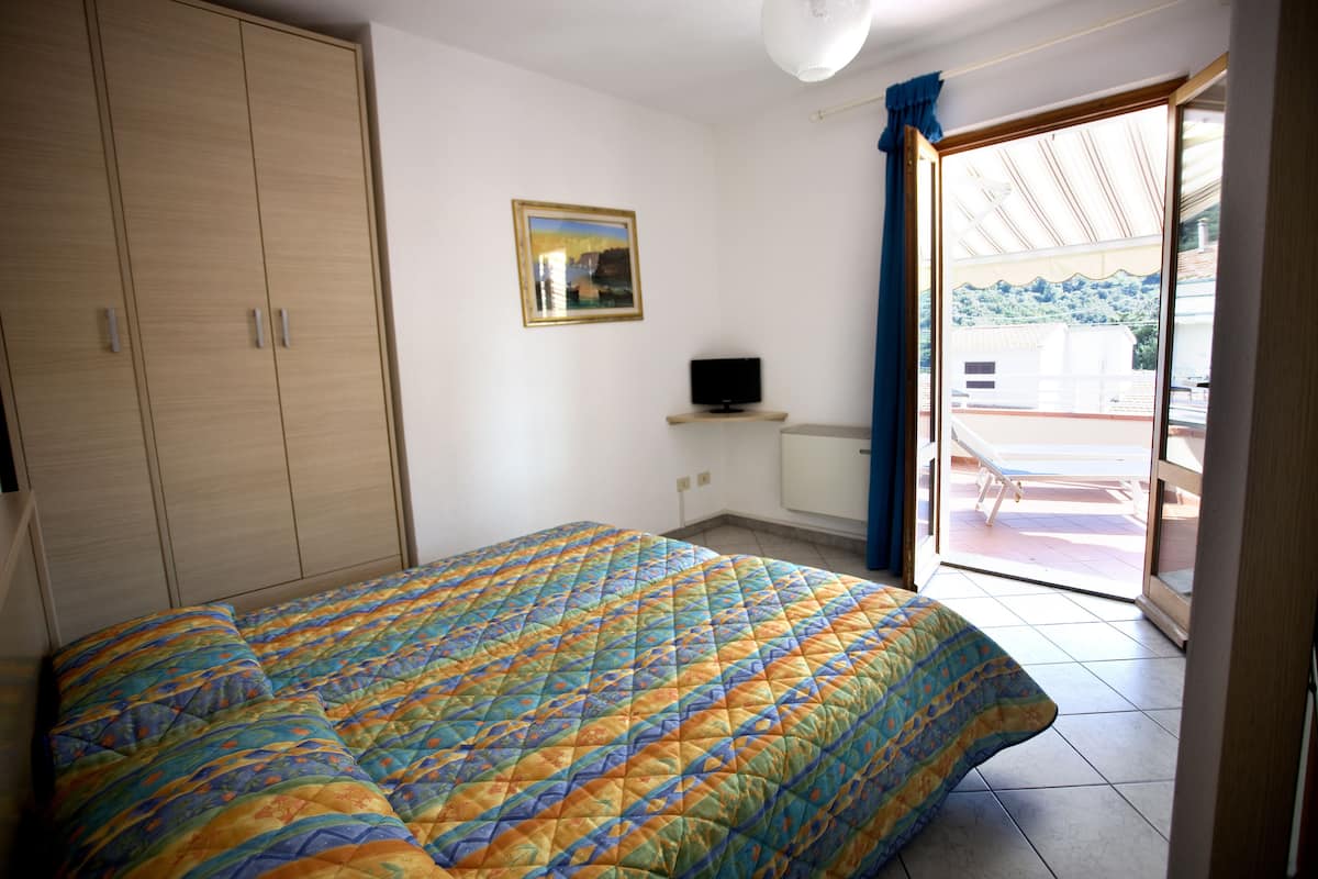 A bedroom with a bed, a TV on a stand, a wardrobe, and a balcony with a view.
