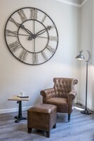 A room with a large wall clock, a leather armchair, a matching ottoman, a small table with a cup, and a floor lamp.