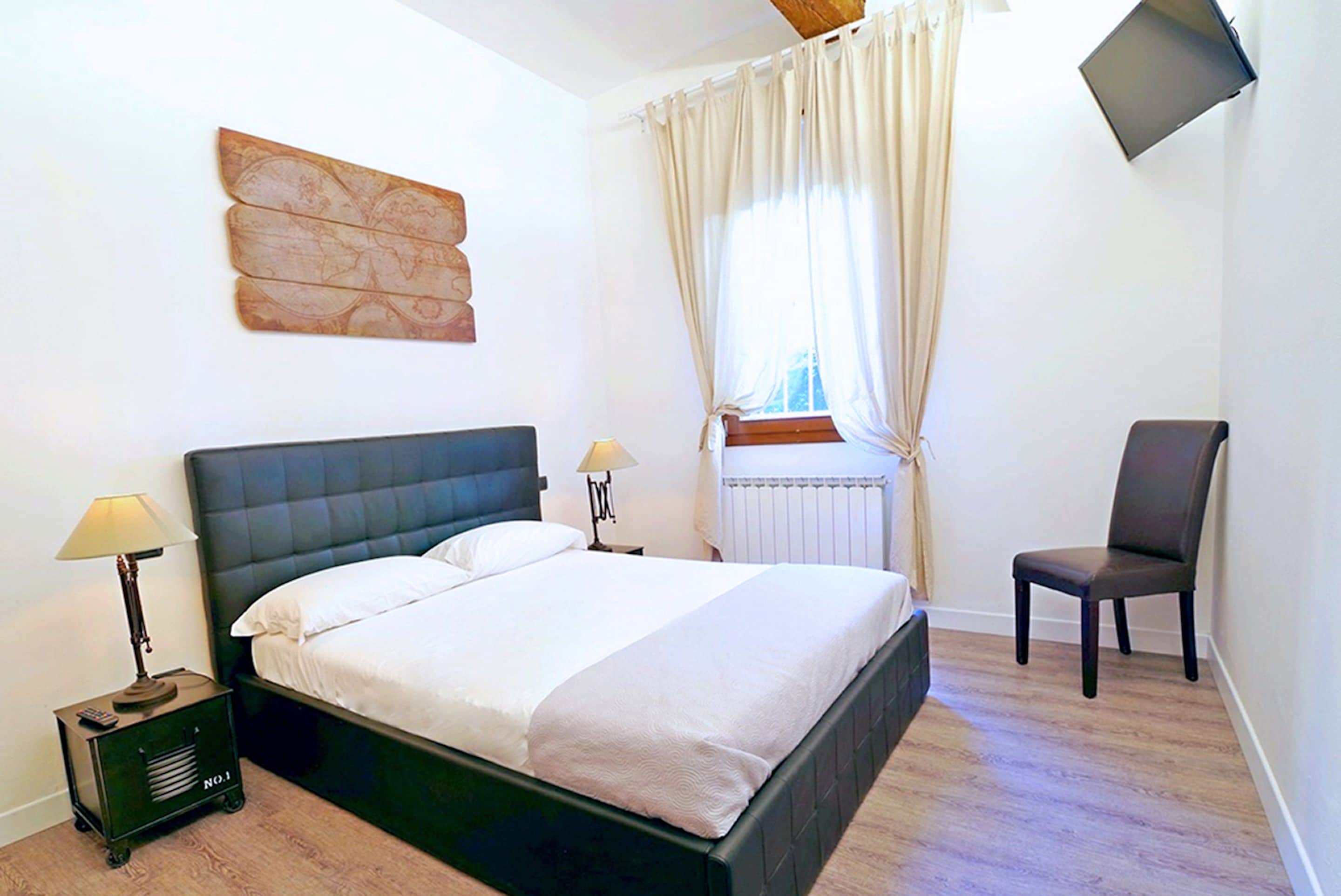 Un dormitorio con cama, dos lámparas de noche, una silla, televisor y ventana con cortinas.