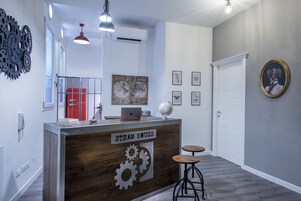 A reception desk labeled "STEAM HOUSE" with a laptop, globe, and framed pictures on the wall. There are two stools and a red cabinet in the background.