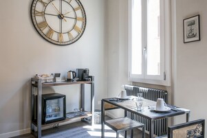 A modern kitchen with a large wall clock, a built-in refrigerator, a coffee machine, and a dining area with a table and chairs.