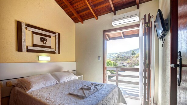 A bedroom with a bed, a wall-mounted TV, and a balcony with a view. - Morada do Sol Apart Hotel Garopaba (Garopaba)