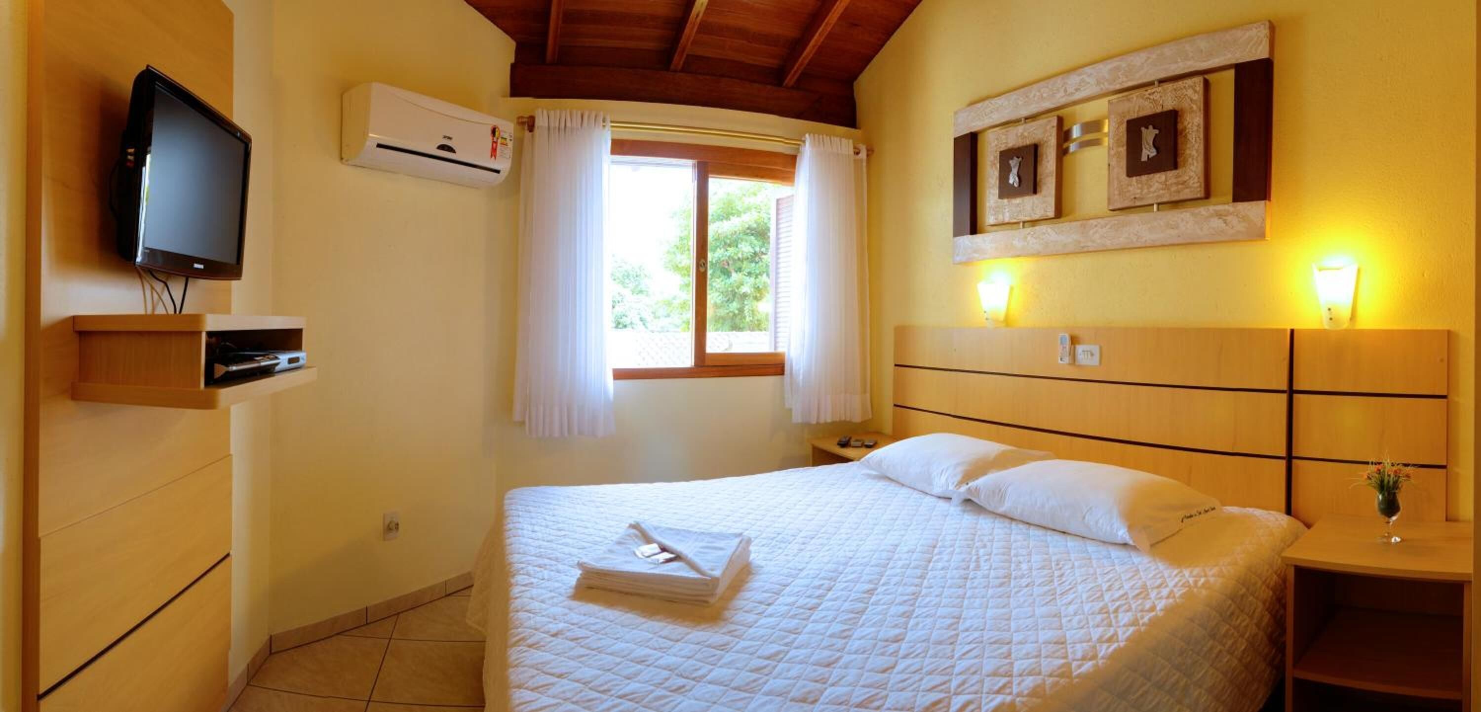 A hotel room with a bed, a TV, an air conditioner, a window with curtains, a bedside table with a lamp, and a wall-mounted picture frame.