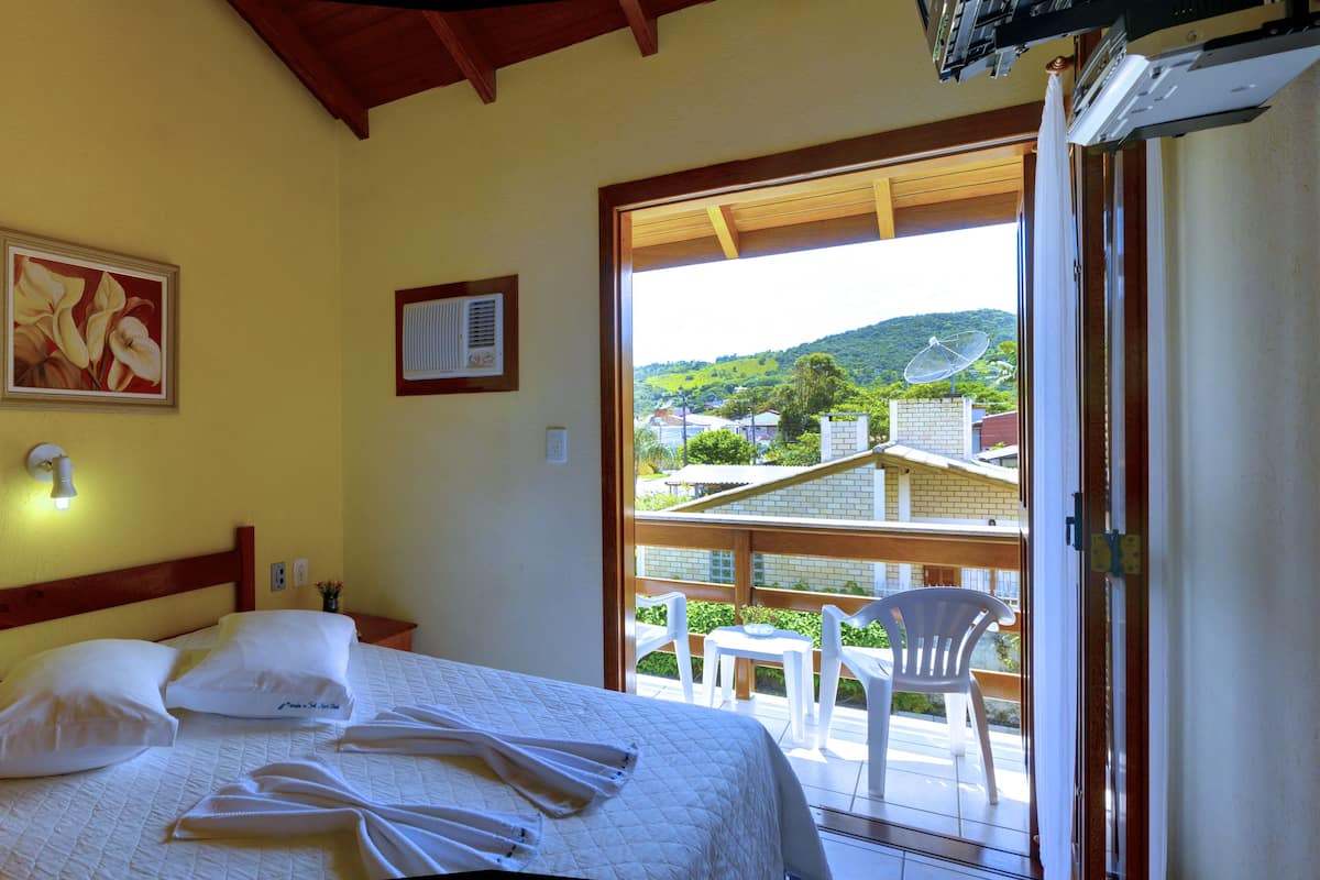 A hotel room with a bed, a painting, an air conditioning unit, and a balcony with a view of the countryside.