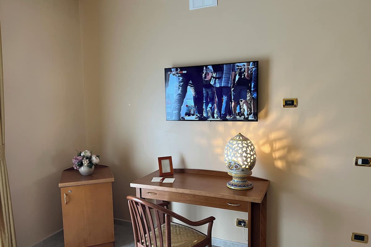 Una habitación con escritorio de madera, una silla, televisor montado en la pared, un armario y una lámpara decorativa.