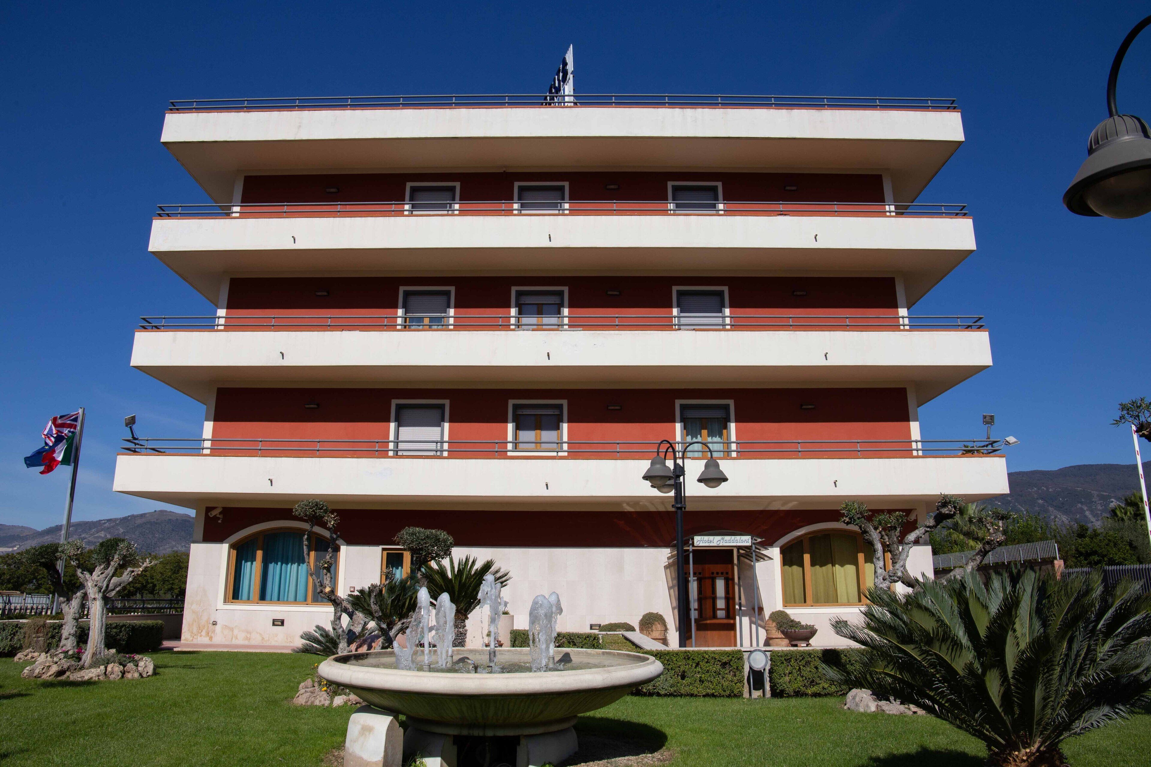 A multi-story hotel with a red and white facade, balconies, and a fountain in the front garden.