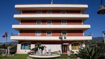 A multi-story hotel with a red and white facade, balconies, and a fountain in the front garden.