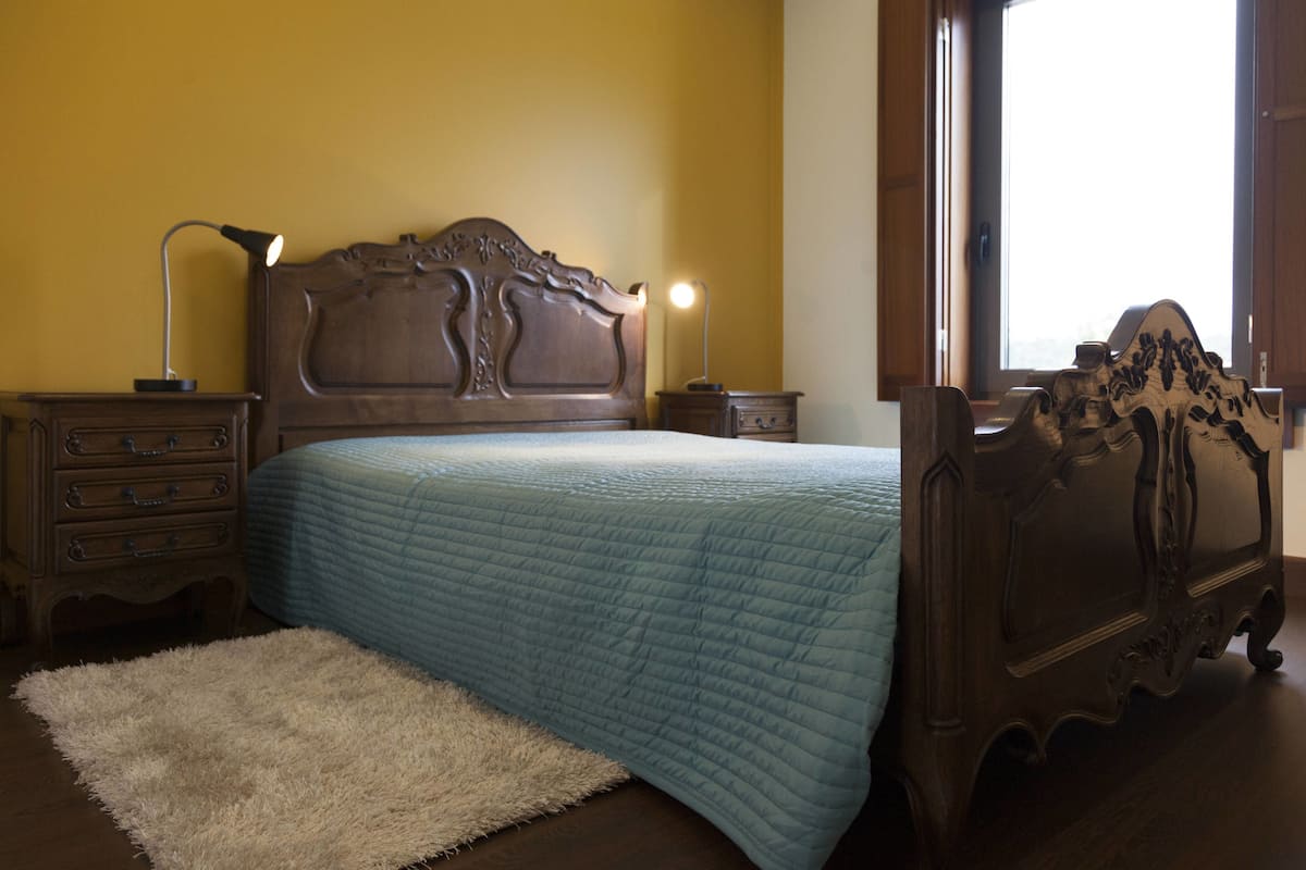 A bedroom with a wooden bed, two bedside tables with lamps, a fluffy rug, and a window with curtains.