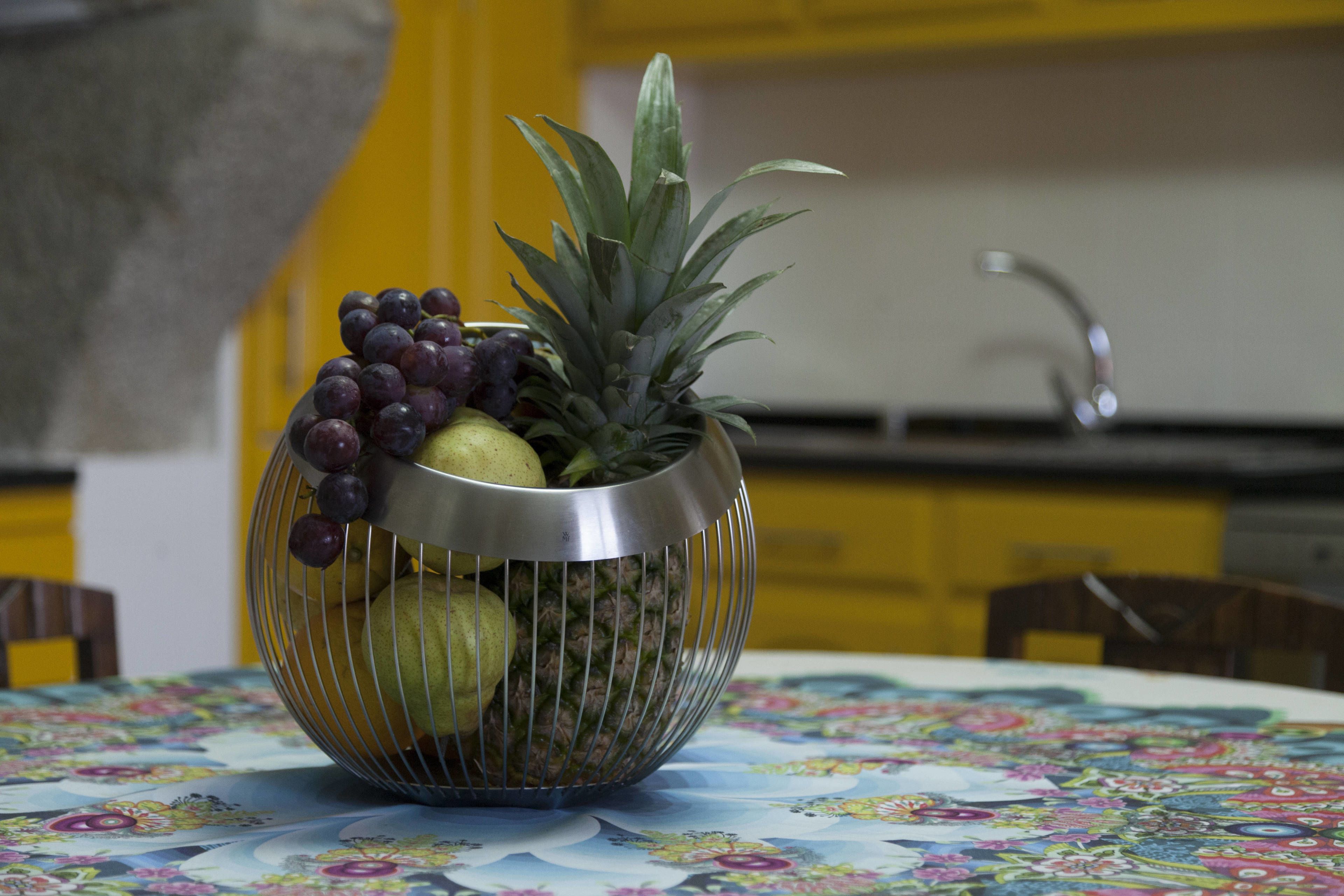 Una cesta de frutas con uvas, piña y peras sobre una mesa con mantel floreado.