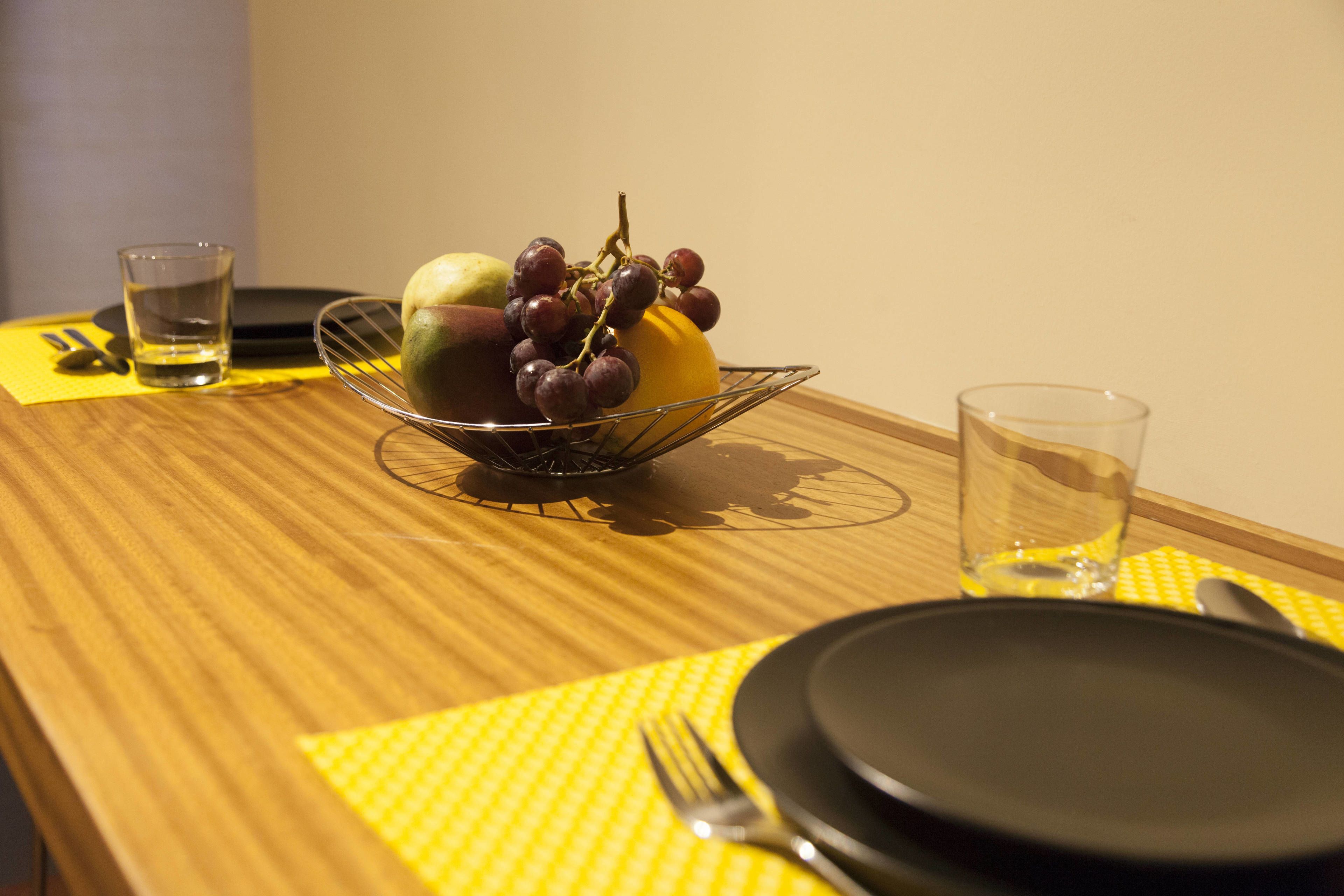 A dining table with a fruit bowl containing grapes and a lemon, a glass of water, and a set of cutlery.