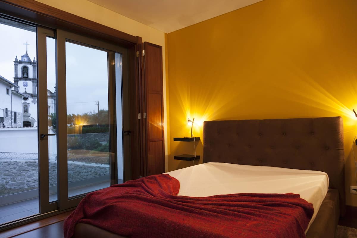 A bedroom with a bed, a nightstand, and a window with a view of a church and street.