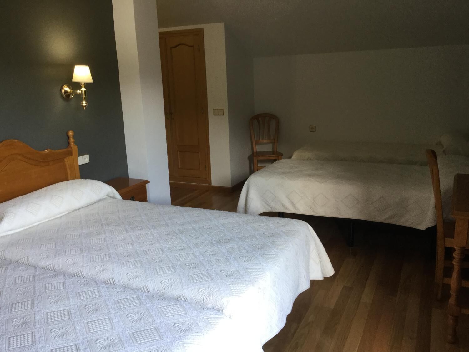 A room with two beds, a wooden headboard, a nightstand, a chair, and a wall-mounted lamp.