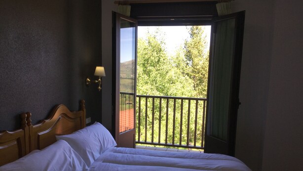Un dormitorio con una cama grande, una mesita de noche, una lámpara de pared y un balcón con vistas a árboles y un edificio de techo rojo.