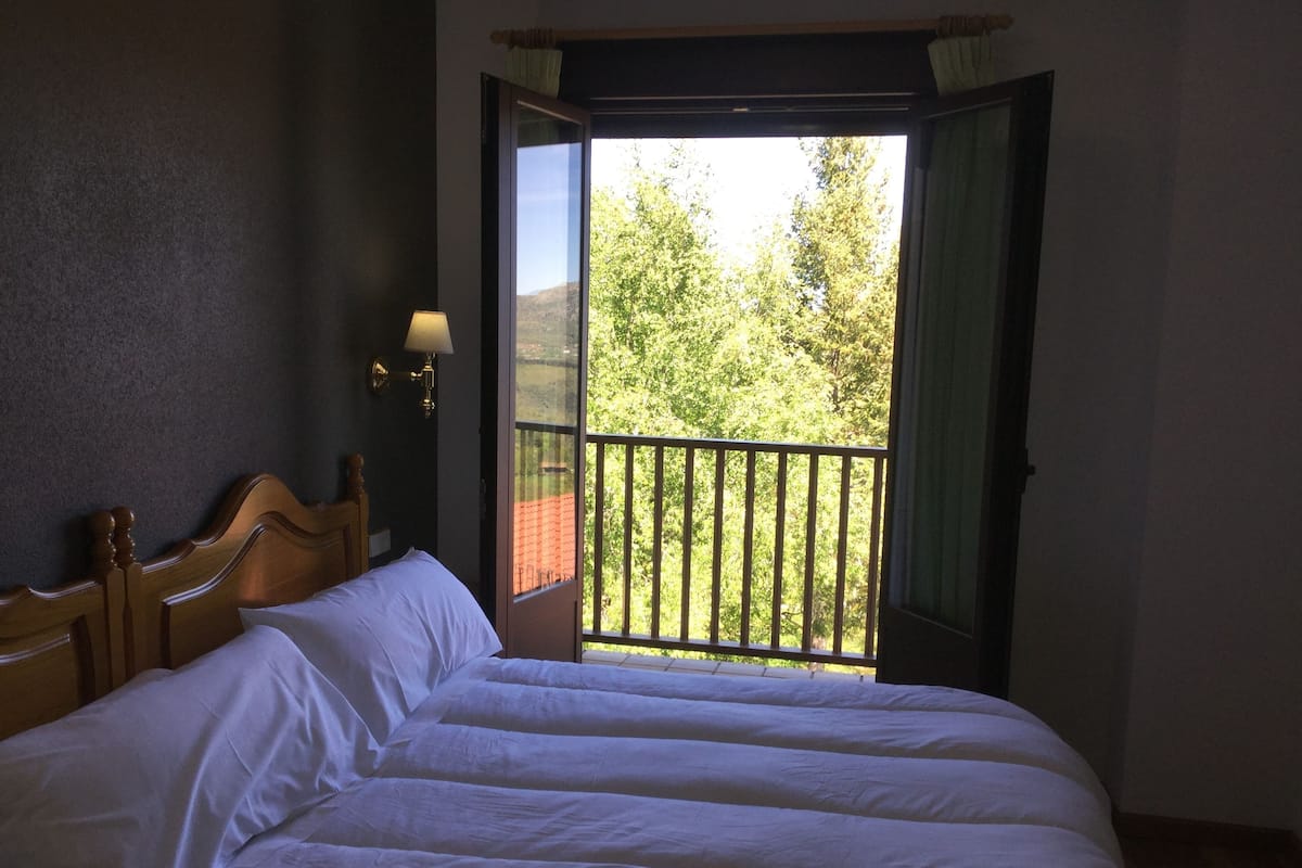 Ein Schlafzimmer mit einem großen Bett, einem Nachttisch, einer Wandlampe und einem Balkon mit Blick auf Bäume und ein Gebäude mit rotem Dach.