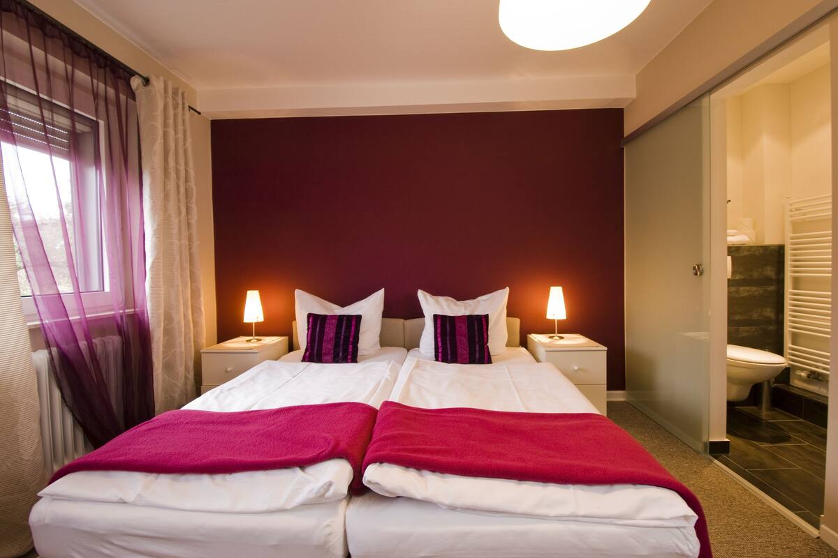 A double bed with white and red bedding, two bedside lamps, and a view of a bathroom.