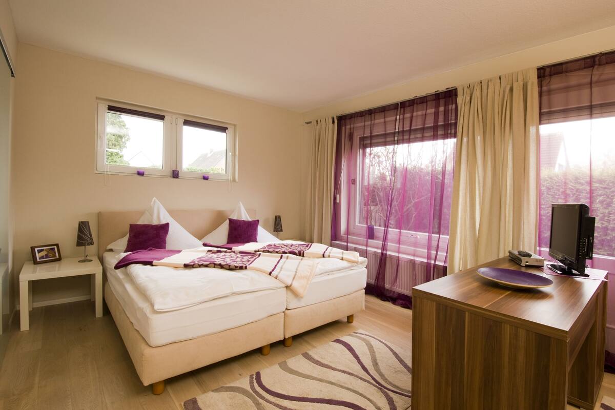 A bedroom with a bed, bedside tables, a desk, a television, and a window with curtains.