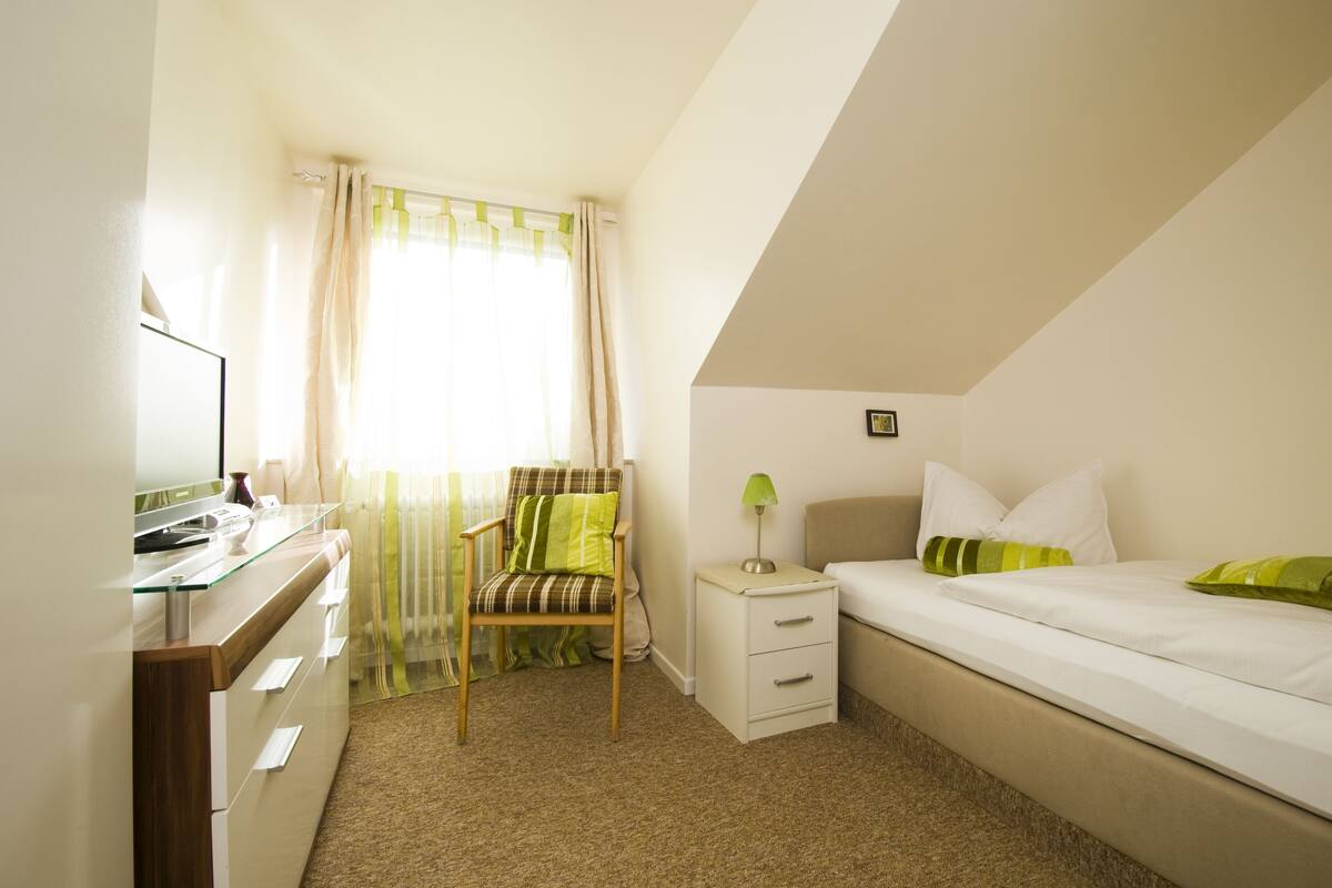 A bedroom with a bed, a chair, a nightstand, a lamp, and a window with curtains.