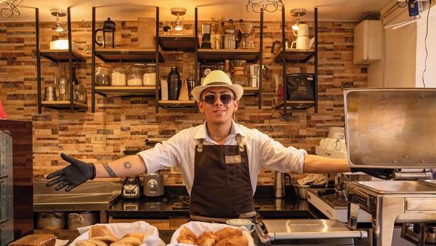 A kitchen with a brick wall, shelves with kitchenware, and a person in a chef's outfit with arms outstretched. - Mayu Boutique Hotel (Cusco)