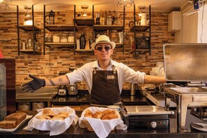 Una cocina con pared de ladrillo, estantes con utensilios de cocina y una persona con uniforme de chef, brazos extendidos.