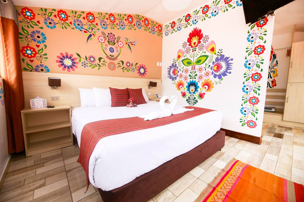 Une chambre d’hôtel avec un grand lit, une table de chevet, un mur décoré de motifs floraux et un plancher en bois.